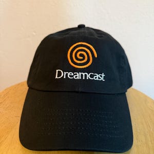 May include: Black baseball cap featuring the Dreamcast logo in orange and the word "Dreamcast" in white. The cap has a curved brim and a classic six-panel design. A stylish accessory for fans of the classic video game console.