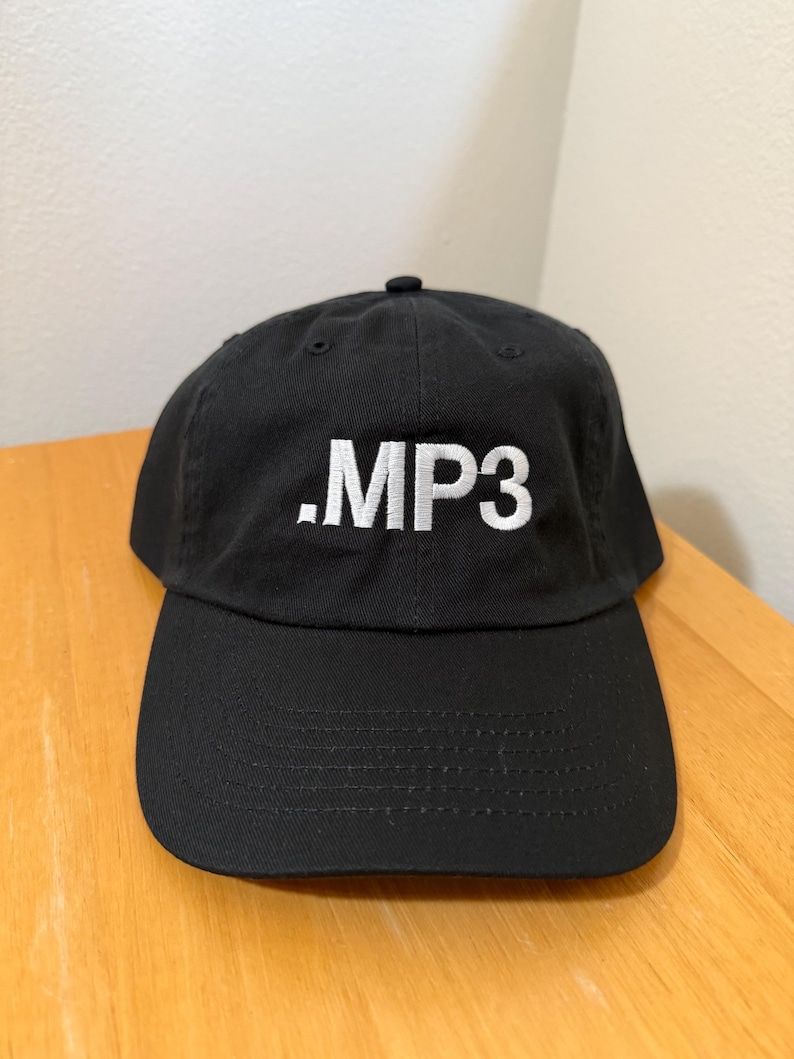 May include: Black baseball cap with a curved brim. The cap features the white text ".MP3" embroidered on the front. The hat is made of a durable fabric and has an adjustable strap at the back.