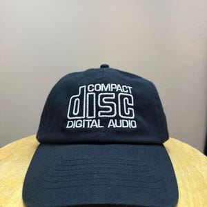 May include: Black baseball cap with the words "COMPACT DISC DIGITAL AUDIO" embroidered in white. The hat has a curved brim and a rounded top.