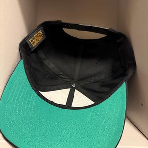 May include: A black baseball cap with a green brim. The interior of the cap is black, and the brim is a vibrant green color. The cap has a snapback closure for adjustable sizing. The label reads "The Classics".