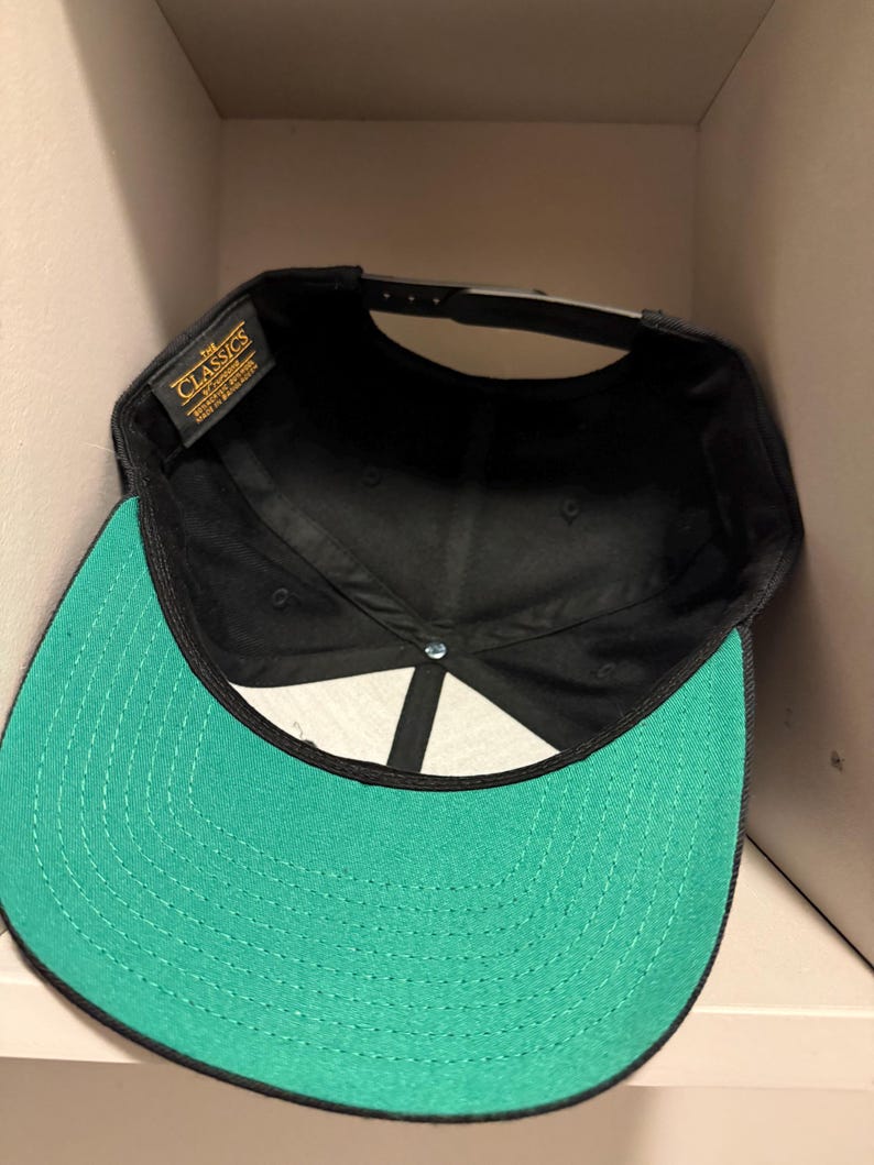 May include: A black baseball cap with a green brim. The interior of the cap is black, and the brim is a vibrant green color. The cap has a snapback closure for adjustable sizing. The label reads "The Classics".