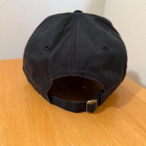 May include: A black baseball cap with a curved brim. The cap has a brass-colored adjustable buckle closure on the back. The cap is made of a solid, dark fabric.