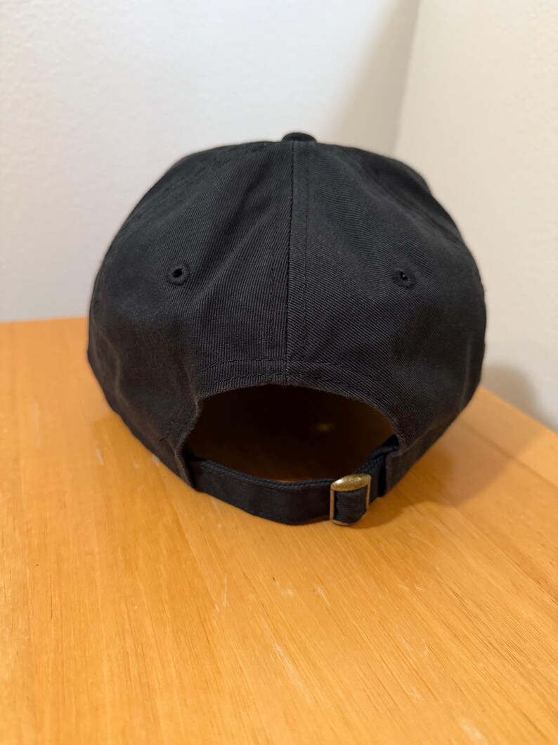 May include: A black baseball cap with a curved brim. The cap has a brass-colored adjustable buckle closure on the back. The cap is made of a solid, dark fabric.