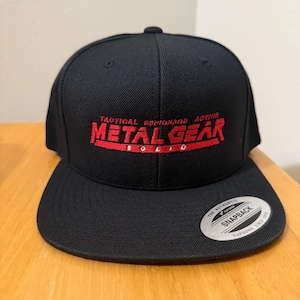 以下が含まれることがあります： 黒いフラットブリムのスナップバックキャップ。赤い文字で「METAL GEAR SOLID」と「TACTICAL ESPIONAGE ACTION」が刺繍されています。背面にはスナップ留めがあり、「THE AUTHENTIC SNAPBACK」ステッカーが付いています。