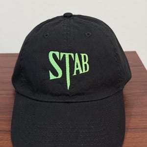 以下が含まれることがあります： フロントパネルに明るいライムグリーンの糸で「STAB」という文字が刺繍された黒い野球帽。この帽子は、構造化されたクラウン、カーブしたつば、上部のボタンが特徴で、シンプルで洗練されたデザインに仕上がっています。