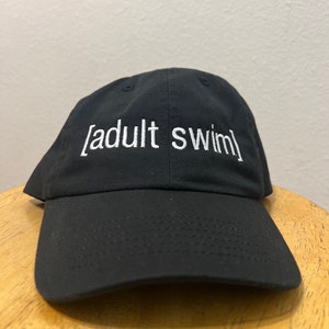 May include: A black baseball cap with white embroidered text that reads "[adult swim]"
