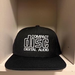 May include: Black baseball cap with a flat brim. The cap features the words "COMPACT DISC DIGITAL AUDIO" in white, stylized text. The hat is made of a textured fabric and is displayed in a neutral-toned shelf.