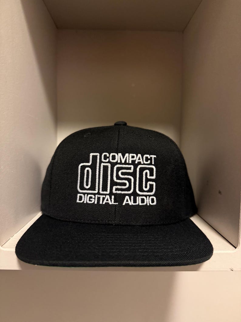 May include: Black baseball cap with a flat brim. The cap features the words "COMPACT DISC DIGITAL AUDIO" in white, stylized text. The hat is made of a textured fabric and is displayed in a neutral-toned shelf.