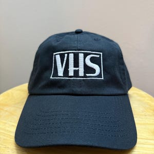 以下が含まれることがあります： 白い長方形の中に白いブロック体で「VHS」と刺繍された黒い野球帽。帽子には湾曲したつばと丸みを帯びた上部があります。
