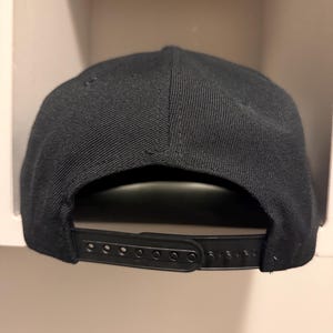 May include: A black snapback baseball cap. The cap features a structured crown and a flat brim. The adjustable strap on the back allows for a customized fit. The cap is made of a durable fabric.