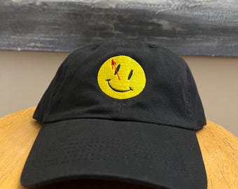 May include: Black baseball cap with a yellow smiley face embroidered on the front. The smiley face has a red slash through it and a red arrow pointing up.