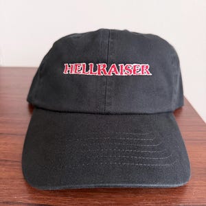 May include: A black baseball cap featuring the word HELLRAISER embroidered in red thread with a white outline on the front panel. The cap has a structured crown, a curved brim with horizontal stitching, and a classic six-panel design construction.
