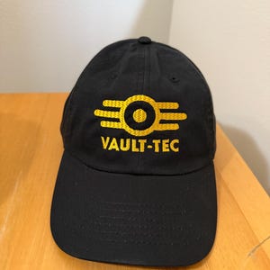May include: Black baseball cap featuring the Vault-Tec logo in gold. The logo is a stylized design with a circle in the center and three horizontal lines on each side. The text "VAULT-TEC" is embroidered in gold below the logo.