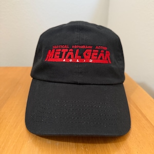 May include: Black baseball cap with the text "TACTICAL ESPIONAGE ACTION METAL GEAR SOLID" embroidered in red. The cap has a curved brim and a classic six-panel design.