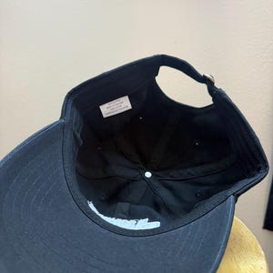 K&ouml;nnte beinhalten: Schwarze Baseballkappe mit gebogenem Schirm und verstellbarem Riemen. Die Unterseite der Kappe zeigt wei&szlig;en Text und ein kleines Etikett mit Text. Die Kappe ist aus Baumwolle.