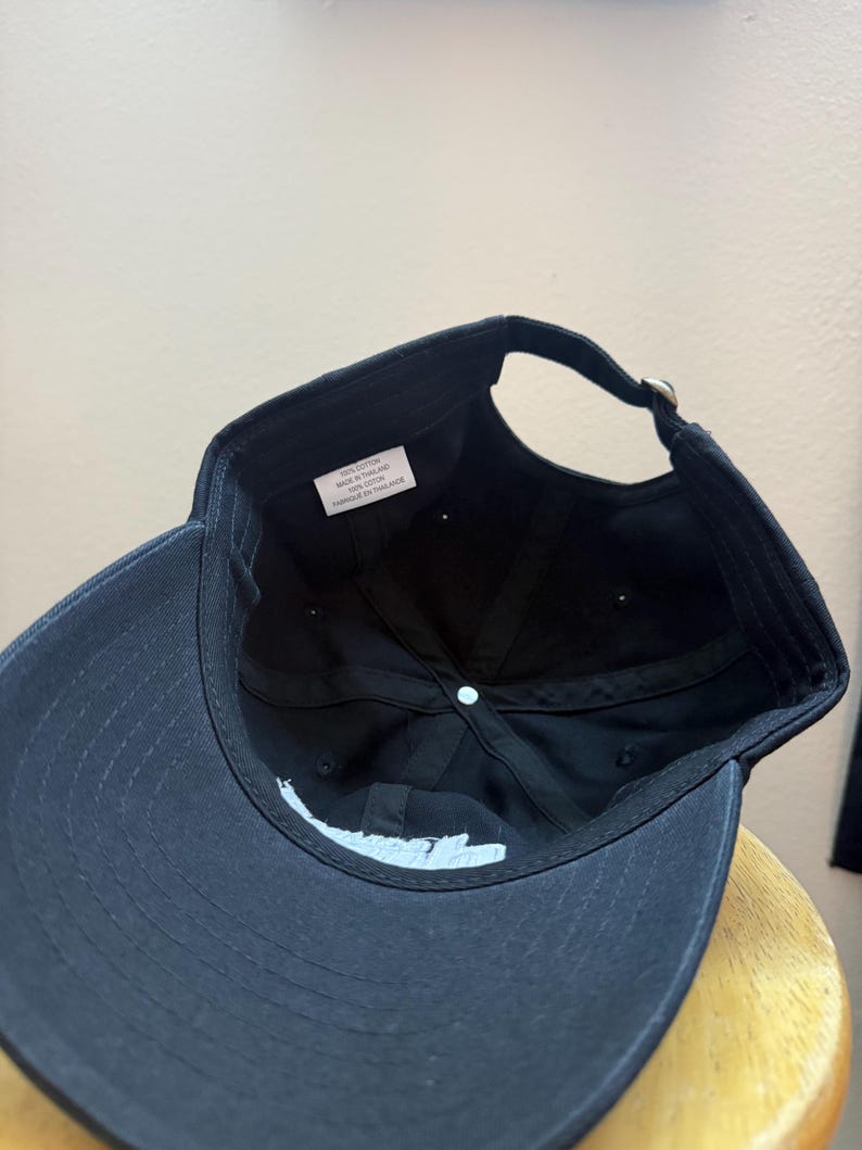 K&ouml;nnte beinhalten: Schwarze Baseballkappe mit gebogenem Schirm und verstellbarem Riemen. Die Unterseite der Kappe zeigt wei&szlig;en Text und ein kleines Etikett mit Text. Die Kappe ist aus Baumwolle.