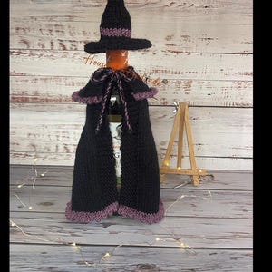 May include: A knitted black and purple bottle cover set consisting of a witch hat topper and a matching cape with a tie closure. The set is designed to fit over a bottle, featuring a dark black base color with purple trim accents on the hat brim, the cape edges, and the pom-pom topper.