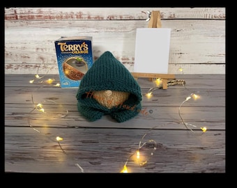 May include: A small, hand-knitted dark teal gnome with a pointed hood and a beige face, positioned next to a blue Terry's Chocolate Orange box and a miniature wooden easel holding a blank white card, surrounded by thin, glowing fairy lights.
