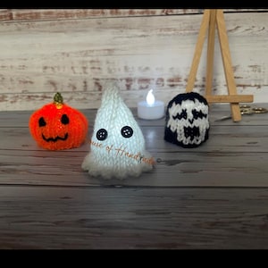 Op de afbeelding: Drie kleine gebreide Halloween-decoraties: een oranje pompoen met een groene steel, een wit spookje met twee zwarte knoopogen en een zwart-wit schedelfiguur. Een klein wit LED-theelichtje en een houten schildersezel staan erachter opgesteld.