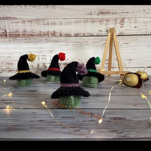 May include: Three small knitted witch hats in black with colourful accents, including yellow, red, purple, and green bands and pom-poms. The hats are placed over green knitted bases, with a wooden easel and gold-wrapped chocolates nearby.