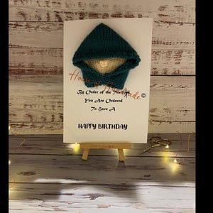Peut inclure: Une carte de vœux blanche avec une petite capuche tricotée bleu sarcelle et un visage crème à l'intérieur. La carte porte le texte : House of Handmade, By Order of the Knitters, You Are Ordered To Have A HAPPY BIRTHDAY, avec un symbole copyright.