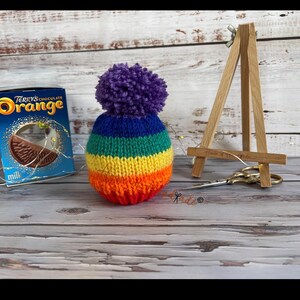 May include: A handmade, colorful knit hat with a purple pom-pom. The hat features bands of rainbow colors: orange, yellow, green, blue, and purple. A box of Terry's Chocolate Orange is visible in the background.