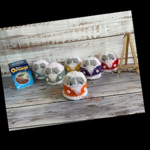 May include: A selection of colourful, knitted miniature camper vans. The vans are in shades of orange, yellow, purple, red, and blue, each with a white roof and grey windscreen. A box of Terry's Orange chocolate is also visible.