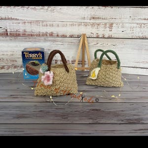 Könnte beinhalten: Zwei kleine, handgefertigte Häkeltaschen in Naturfaserfarbe. Eine Tasche hat braune Griffe und rosa und blaue Blumenverzierungen. Die andere hat grüne Griffe und eine weiße und gelbe Blume. Eine Schachtel Terry's Chocolate Orange befindet sich im Hintergrund.