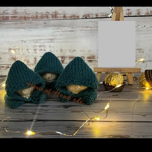 May include: Three small, round objects wrapped in gold foil, each wearing a dark teal knitted hood. The items are arranged near a wooden easel holding a blank white card, accompanied by a string of warm white fairy lights on a grey surface.