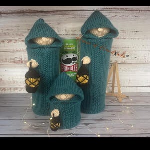 May include: Three teal knitted cozies shaped like hooded figures, each featuring a beige face and holding a small black and yellow lantern. The cozies are placed around a green Pringles can, with the tallest two positioned behind the shortest one.