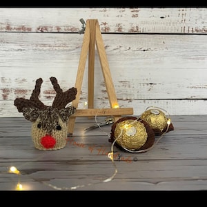 May include: A knitted reindeer head cover with a bright red nose and brown antlers, positioned next to a small wooden easel and two gold-wrapped spherical chocolates. A thin string of small warm white fairy lights is draped around the items.