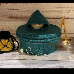 May include: A dark teal knitted round container with a matching lid and a small hood-like top piece. Beside it sits a small yellow and black drawstring pouch with a lattice pattern. The items are displayed with a wooden easel and string lights.