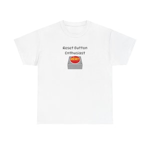 Puede incluir: Camiseta blanca con el texto "Reset Button Enthusiast" y un gráfico de un botón "RESET" rojo en una caja gris. Una prenda casual para el día a día. El diseño es sencillo y llamativo.