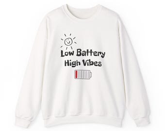 Puede incluir: Sudadera blanca con el texto "Low Battery High Vibes" en negro, un sol sonriente y un icono de batería rojo y blanco. La sudadera tiene cuello redondo y mangas largas.
