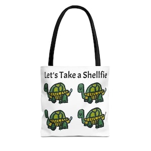 Puede incluir: Bolso tote blanco con asas negras. Presenta el texto "Let's Take a Shellfie" y cuatro tortugas de dibujos animados. Las tortugas son verdes con diseños de caparazón amarillos y marrones. La bolsa está diseñada para transportar objetos.
