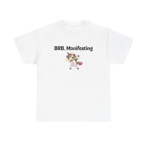 Könnte beinhalten: Weißes T-Shirt mit dem Text "BRB, Manifesting" und einem Cartoon-Einhorn mit Regenbogenmähne und -schwanz, das dabbt. Das Einhorn hat ein goldenes Horn und ist von goldenen Sternen umgeben.
