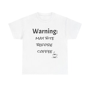 以下が含まれることがあります： 「Warning: MAY BITE BEFORE COFFEE」の黒い文字と小さなコーヒーカップのグラフィックがプリントされた白いTシャツ。カジュアルな普段着です。