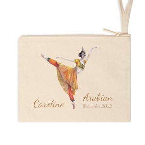 May include: A beige canvas zipper pouch featuring a watercolor illustration of a dancer in an Arabian costume. The dancer is wearing a yellow top, orange pants, and a jeweled headpiece. The text "Caroline Arabian Nutcracker 2022" is printed on the pouch.