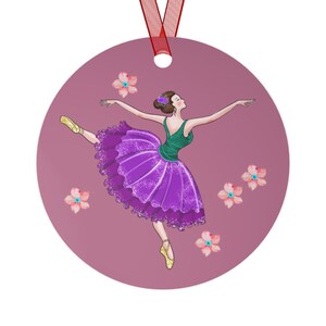 May include: Round ornament featuring a ballerina in a green top and purple tutu, arms outstretched. The illustration includes pink flowers and is set against a mauve background. A red ribbon is attached for hanging.
