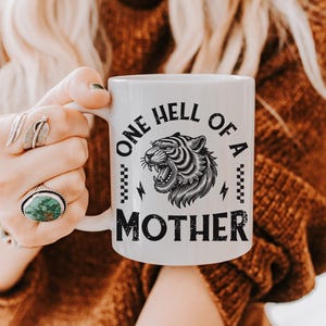 May include: White ceramic mug with a black and white illustration of a tiger and the text "One Hell of a Mother".