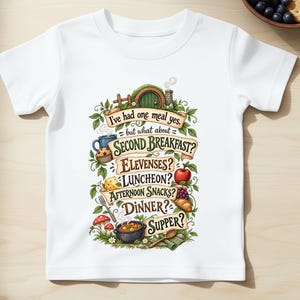 May include: A white short-sleeved t-shirt featuring a colorful graphic print with the text: I've had one meal yes, but what about SECOND BREAKFAST? ELEVENSES? LUNCHEON? AFTERNOON SNACKS? DINNER? SUPPER? surrounded by illustrations of food and a round door.