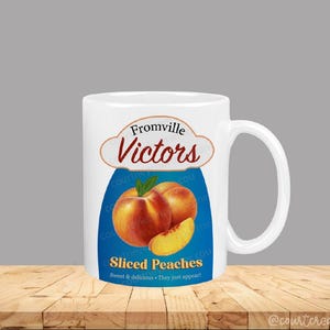 Może przedstawiać: Biały ceramiczny kubek z nadrukiem z napisem "Fromville Victors" i ilustracją brzoskwiń. Projekt zawiera niebieskie tło ze słowami "Sliced Peaches" i "Sweet & delicious. They just appear!"