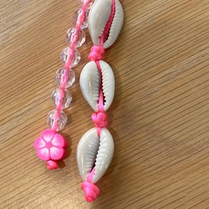 Puede incluir: Un llavero de conchas rosa y blanco con un anillo de metal en forma de corazón. El llavero tiene tres conchas blancas con cuerda rosa y un dije de flor rosa.