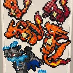 May include: A white magnetic dry-erase board by U Brands featuring five pixel art figures made from colorful plastic beads. The designs include four orange and yellow fire-type creatures and one dark gray and blue creature with wings and a tail.