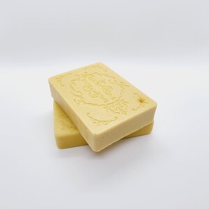 May include: Two bars of pale yellow soap stacked on top of each other. The top bar has a decorative design embossed on the top.