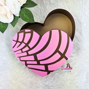 May include: A pink and brown heart-shaped box with a decorative pattern. The box is open and the inside is brown. The box is on a white fluffy surface.