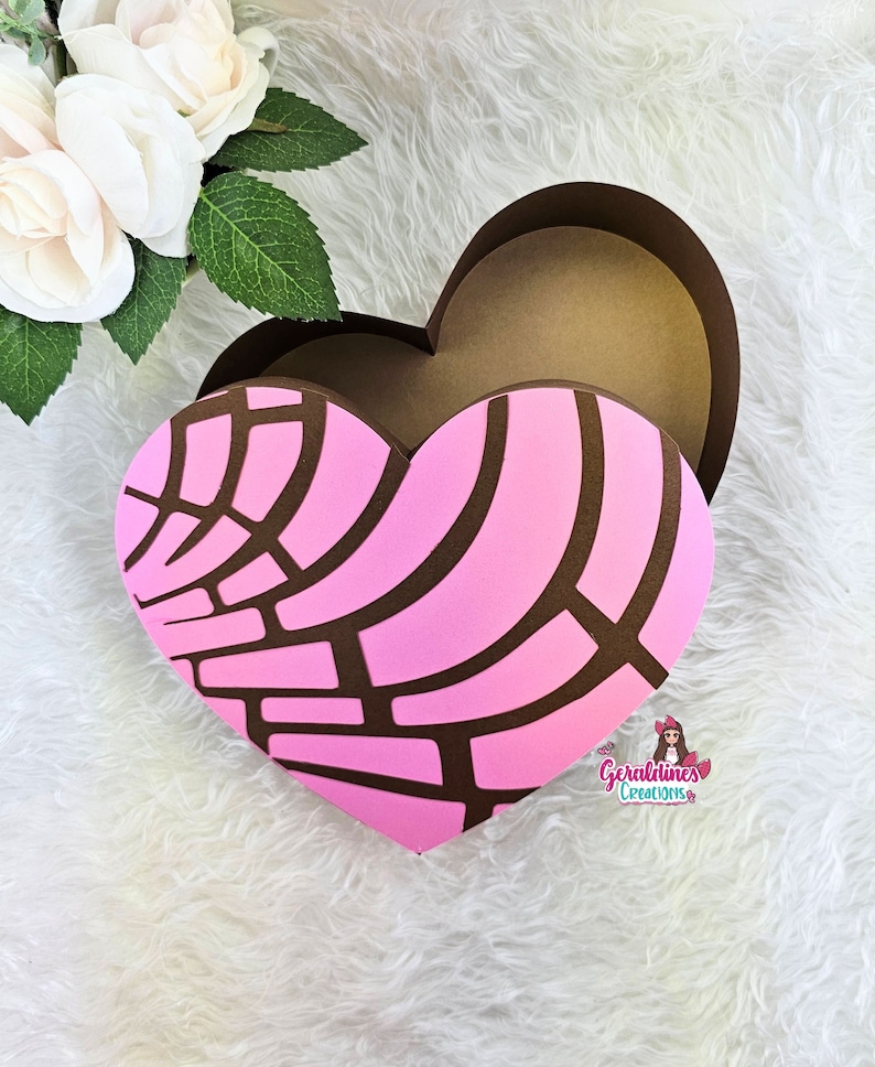 May include: A pink and brown heart-shaped box with a decorative pattern. The box is open and the inside is brown. The box is on a white fluffy surface.