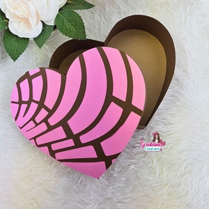 May include: A pink and brown heart-shaped box with a decorative pattern. The box is open and the inside is brown. The box is on a white fluffy surface.
