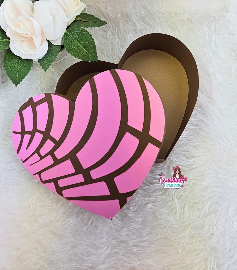 May include: A pink and brown heart-shaped box with a decorative pattern. The box is open and the inside is brown. The box is on a white fluffy surface.