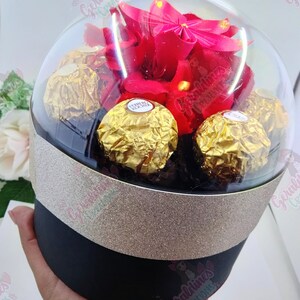 Może przedstawiać: Przezroczysta szklana kopuła z czerwonym kwiatem origami i czekoladkami Ferrero Rocher w środku. Kopuła spoczywa na czarnym cylindrze z złotą wstążką z brokatem.